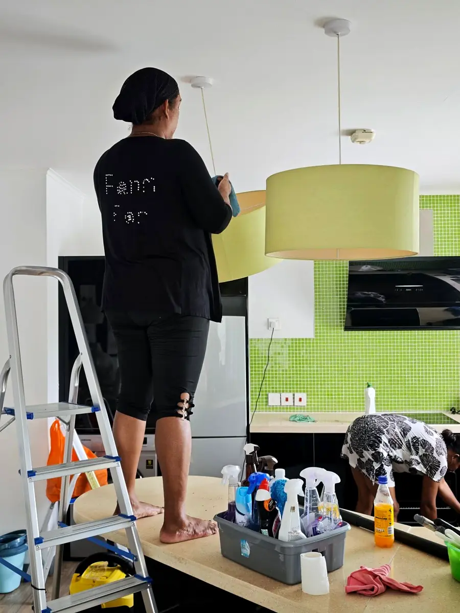 Cleaner dusting a pendant lamp from a ladder in a kitchen