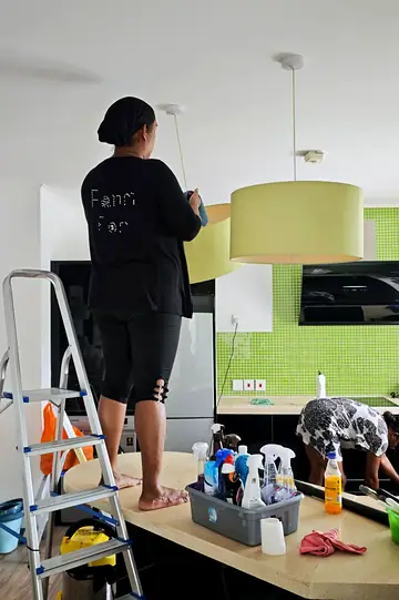 Cleaner dusting a pendant lamp from a ladder in a kitchen